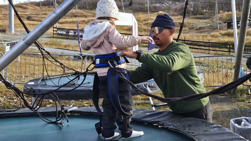 Ansatt i sommerparken fester seler på liten jente, slik at hun kan hoppe på gyro-trampolinen. Hun står oppå trampolinen. Det er høst, hun har på hvit lue. 