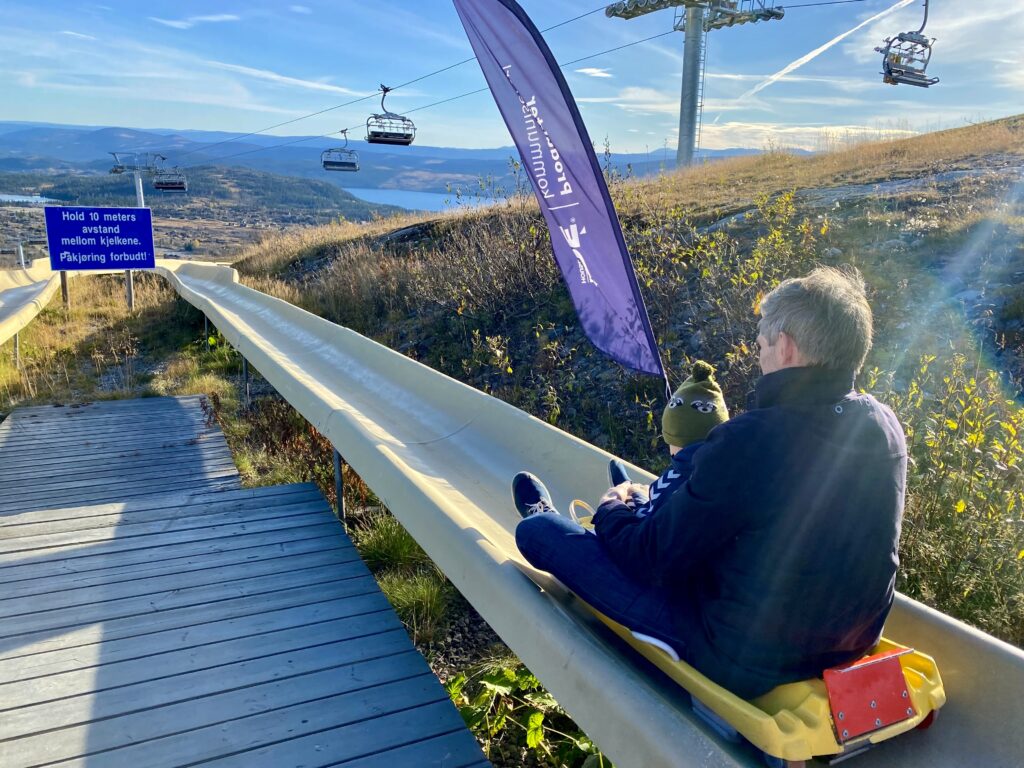 Mann sitter i bob i en bobane, med liten gutt på fanget. På fjellet på Beitostølen. Blå himmel.