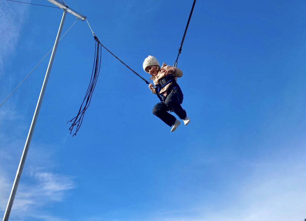 Jente er festet i sele, og flyr høyt i luften. Blå himmel. Hun hopper på gyrotrampoline.