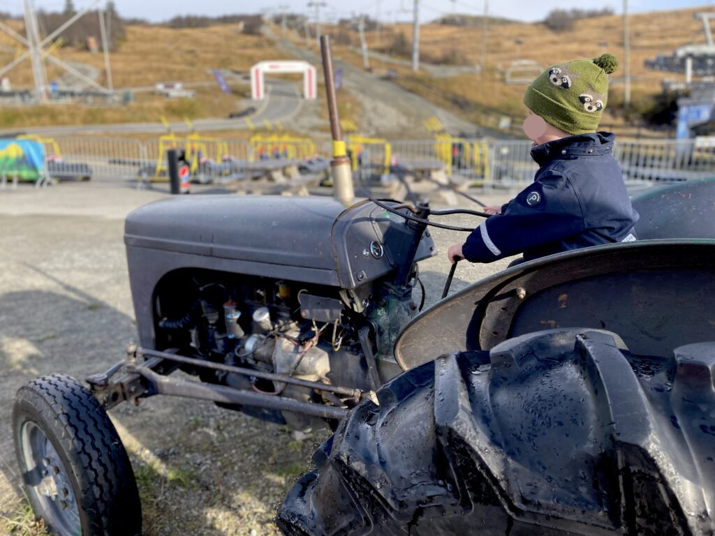 Liten gutt sitter på Gråtass-traktor, med olabilbanen til Beitostølen sommerpark i bakgrunnen. Høst. Gutten har på grønn lue.