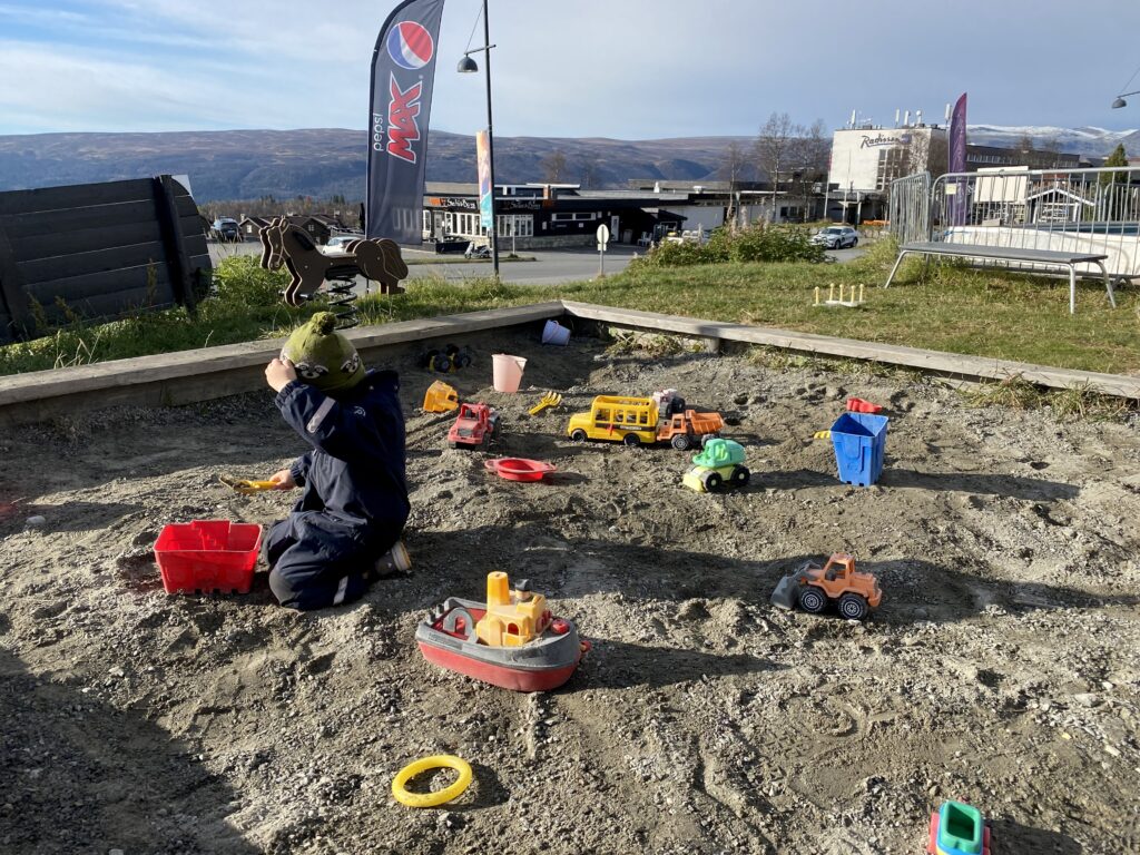 Liten gutt leker i sandkasse. Fjell i bakgrunnen. Sol og høst.