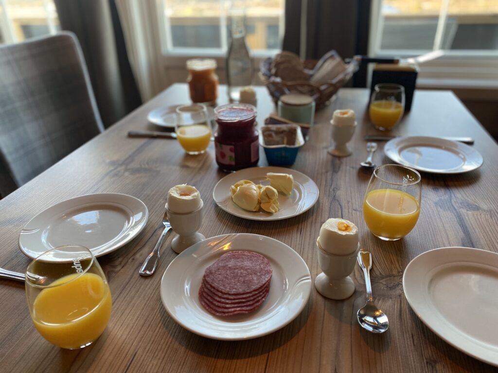 Frokostbord med glass med appelsinjuice, salami, egg i eggeglass, tallerkener, brød, syltetøy.